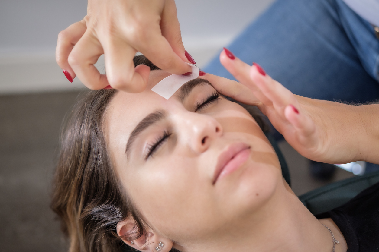 A woman receiving a facial as a technician applies a small adhesive patch to her forehead while she rests with eyes closed.