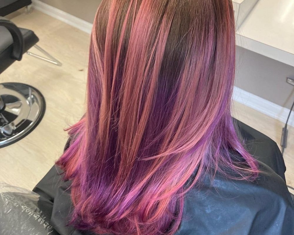 Back view of a person in a salon chair with shoulder-length hair dyed a pink-to-purple gradient.