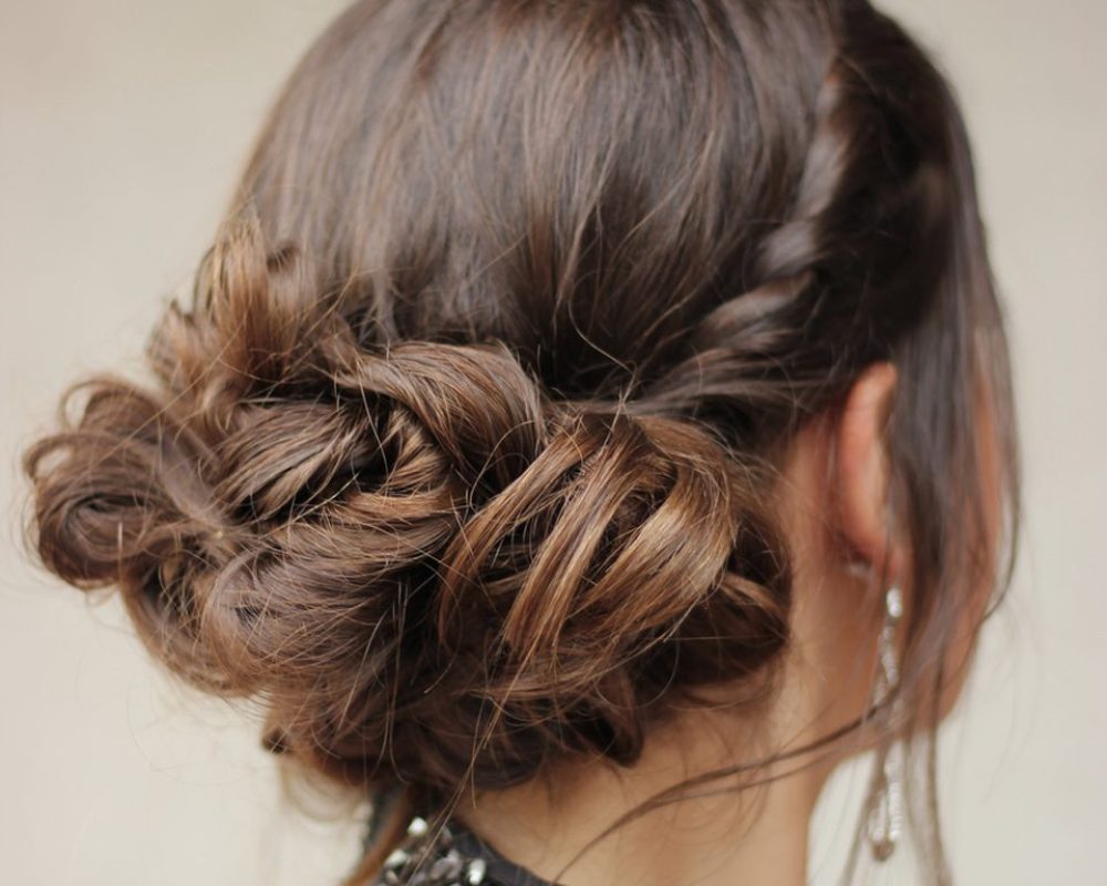 Back view of a brunette braided bun updo, with a sequined black dress and dangling earrings.