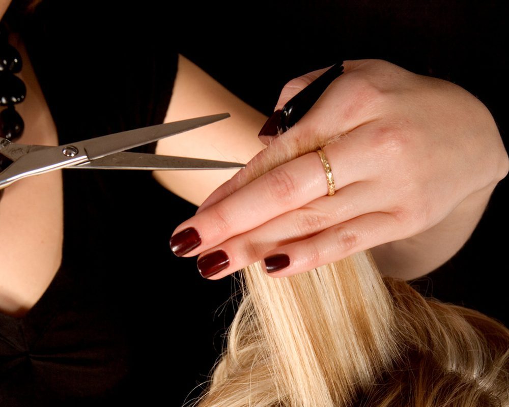 Hairdresser cuts a strand of blonde hair with scissors, fingernails painted dark red, wearing a gold ring, against a black background.