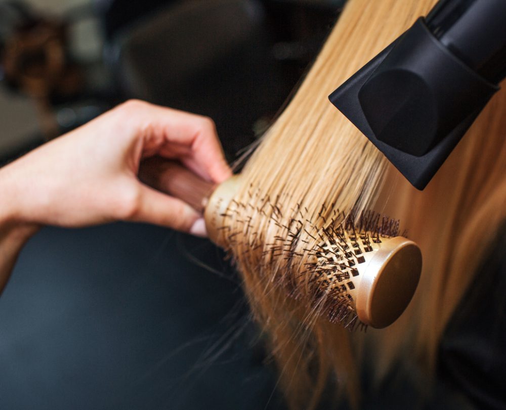 Female client in beauty salon. Close-up of hairdressers hand drying blond hair with hair dryer and round brush, doing professional hairstyle